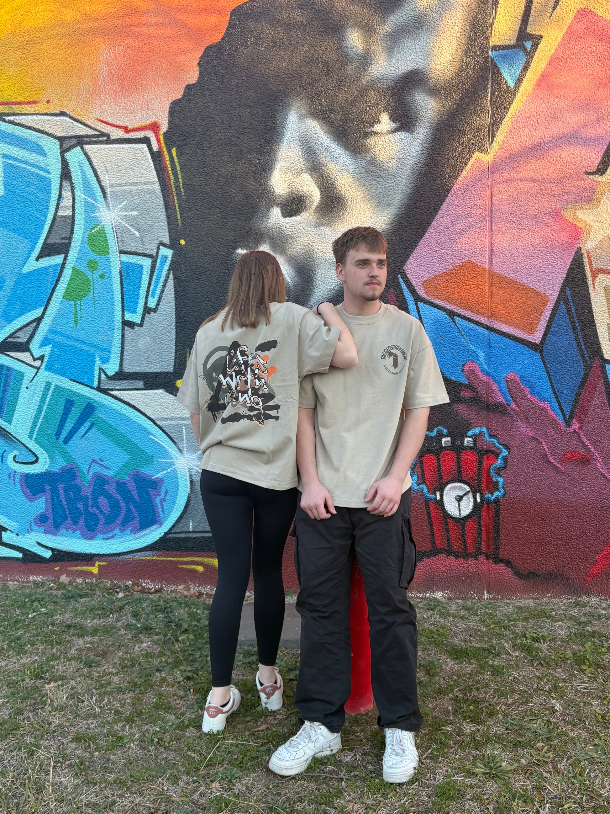 Two people standing in front of a colorful graffiti design in taggedstreetwear 